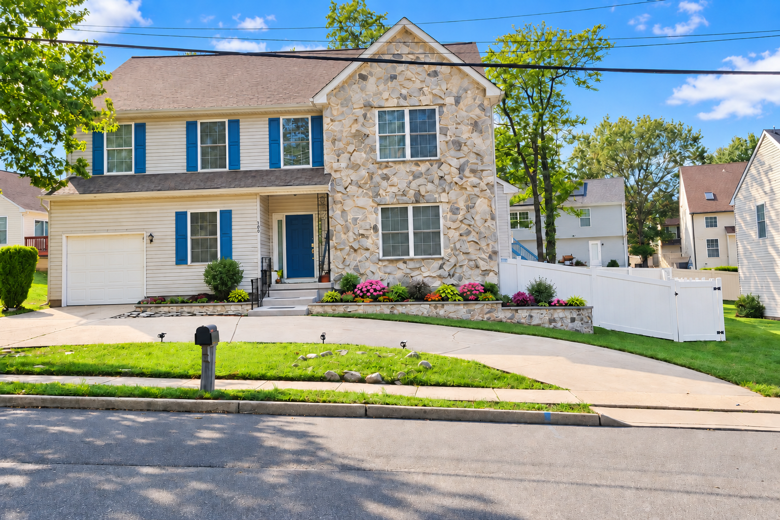 Bright daytime view of the finished front exterior, driveway, retaining wall, flowers, and white fence